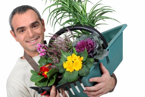 The image depicts a smiling man holding a green plastic garden tool carrier filled with a variety of flowering plants and small shrubs, set against an indoor background. The plants include vibrant yellow, purple, pink, and red blooms, along with lush green foliage. The carrier also contains a small potted plant with long, slender leaves extending upward. The scene highlights gardening and plant care activities, which are relevant to professional gardening services offered in the Willesden area, including hedge trimming and outdoor maintenance. The lighting appears bright and natural, emphasizing the fresh and healthy appearance of the plants, suggesting a focus on garden cultivation and landscaping. The manâ€™s friendly expression and the colorful plants symbolize a well-maintained and vibrant garden environment, reflecting quality garden care services typical of local specialists such as Hedge Trimming Willesden.